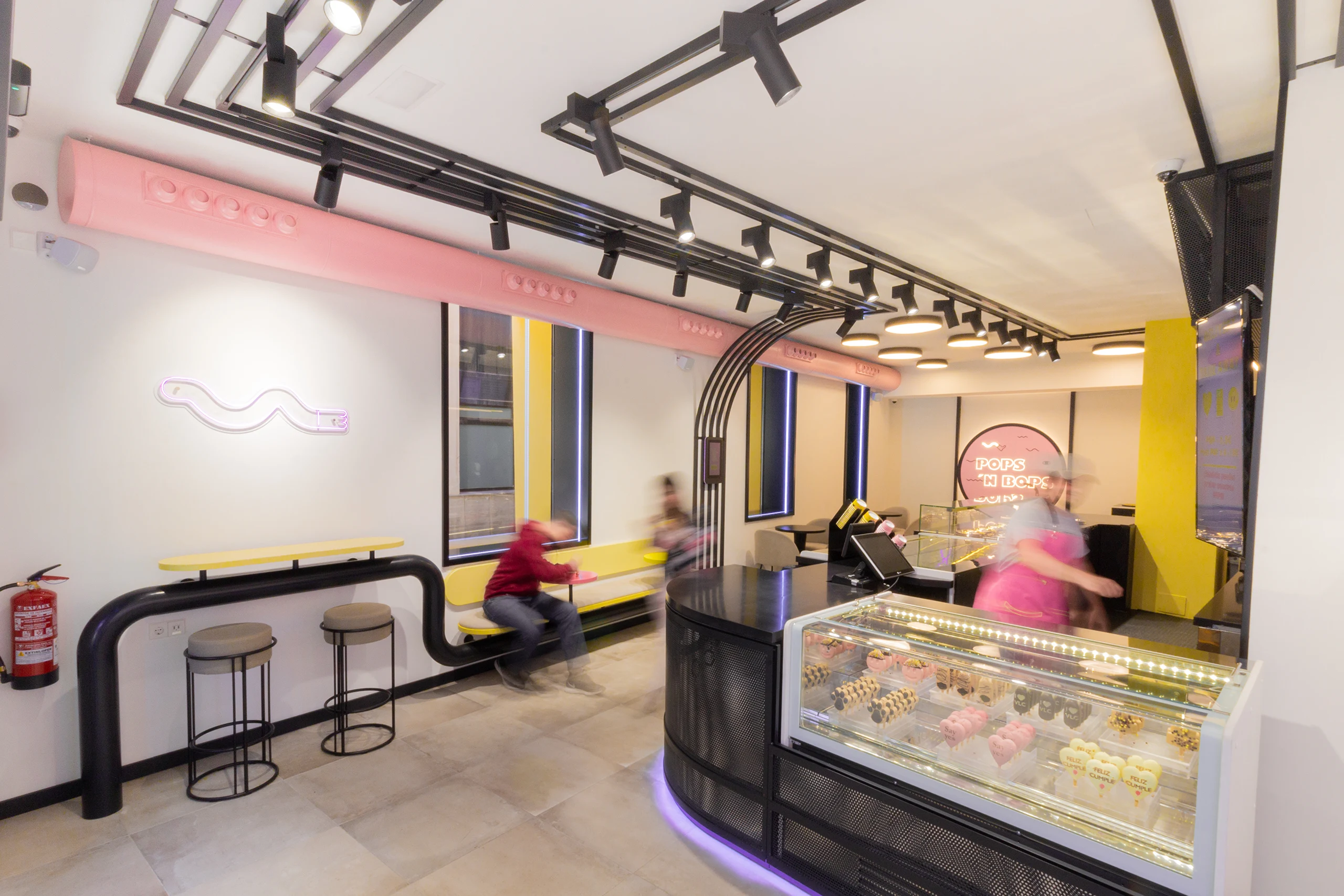 Zona de barra y asientos altos en Pops 'n Bops con tubería rosa, mobiliario amarillo y vitrina expositora de helados