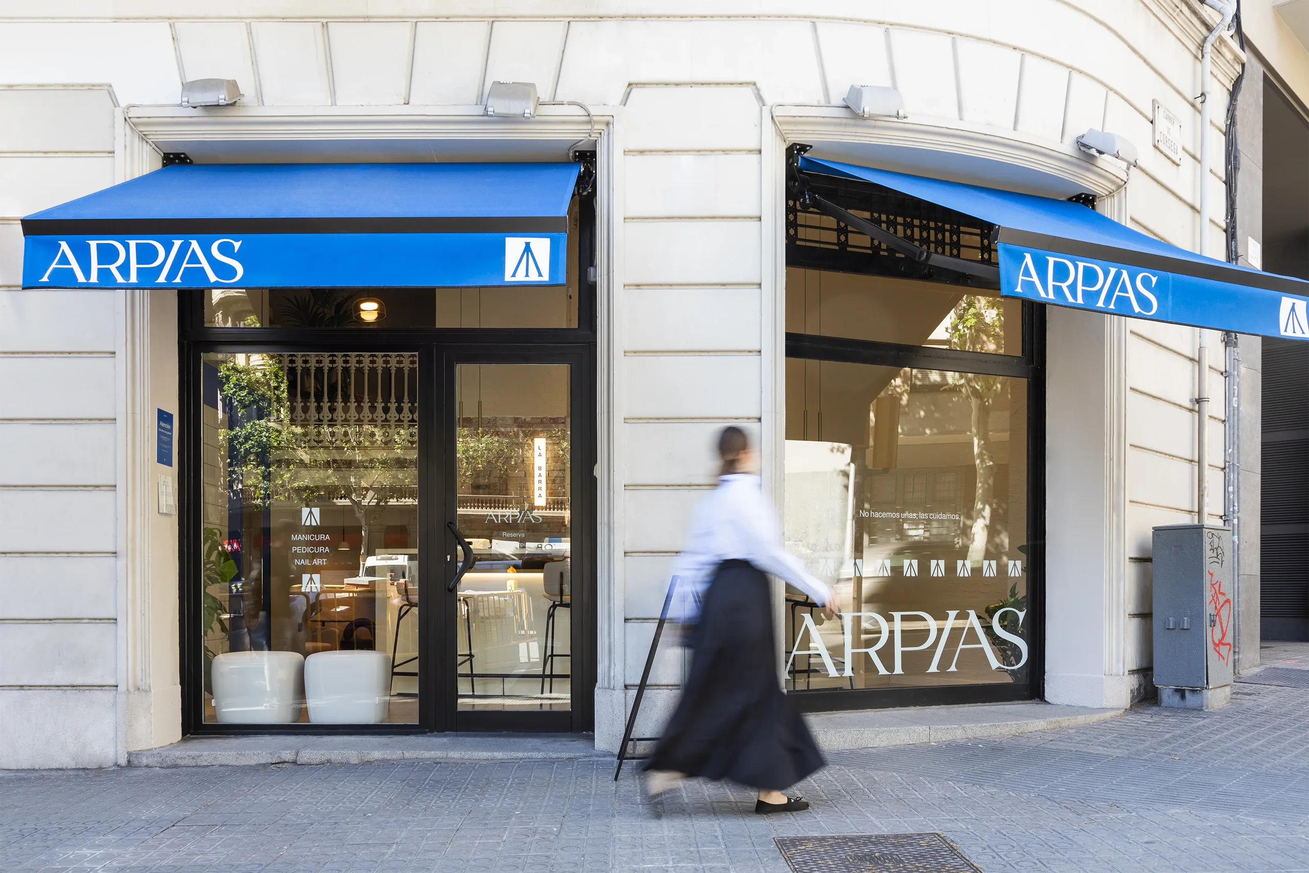 Fachada en chaflán de Arpías en la calle Còrsega con Bailèn (Eixample, Barcelona) con toldos azules, escaparate y vinilos del salón de manicura