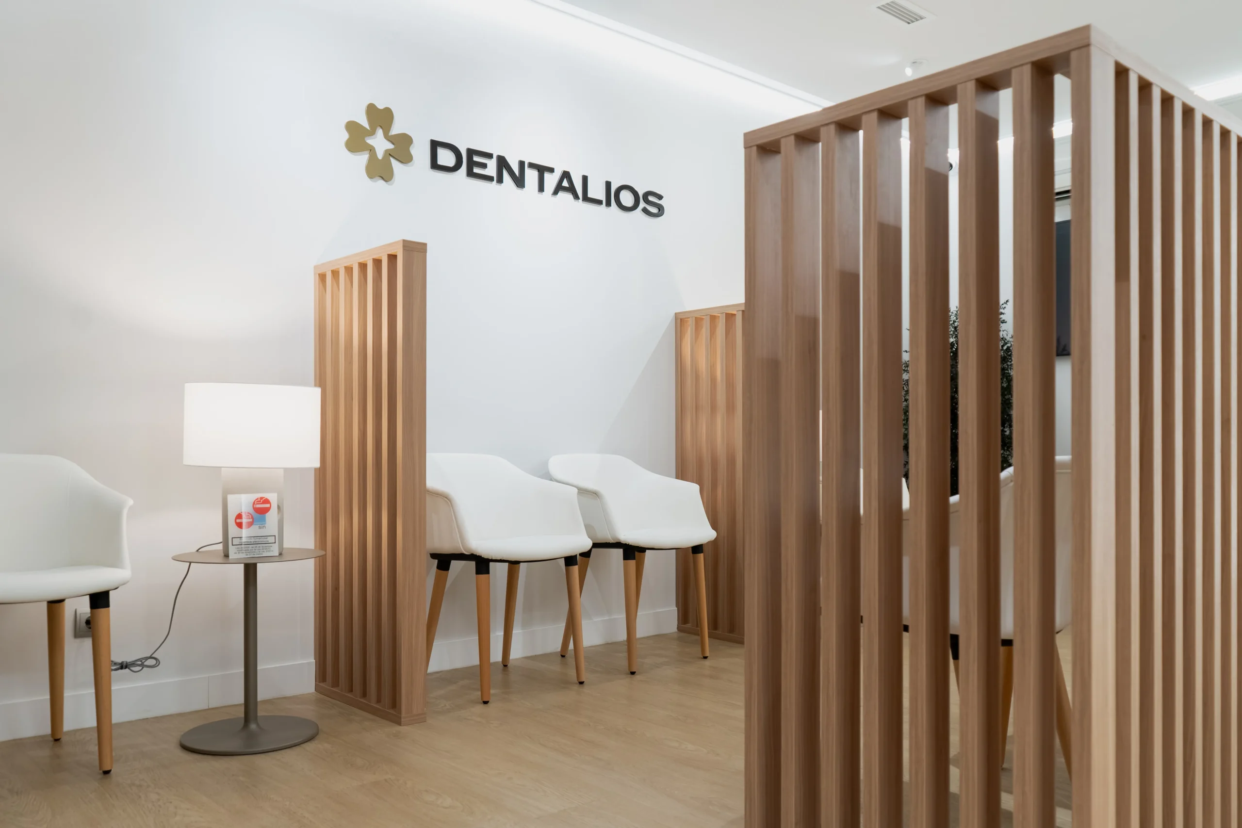 Rincón de espera de Dentalios con sillones blancos, lámpara de sobremesa y el logotipo de la marca sobre pared blanca junto a una celosía de madera