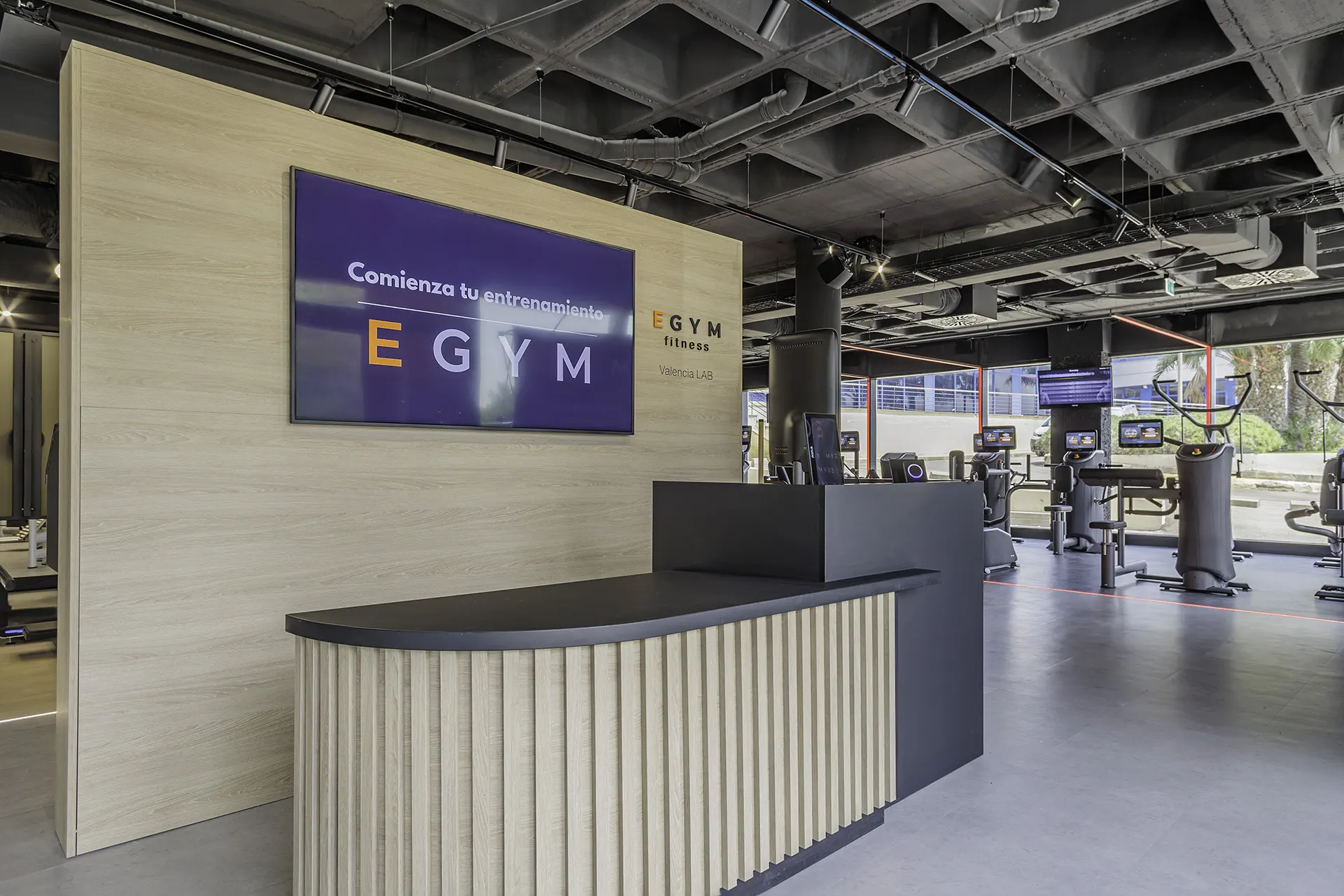Mostrador de recepción de EGYM Fitness Lab con frente de madera acanalada, sobre en negro y pantalla de bienvenida «Comienza tu entrenamiento EGYM», con la zona de cardio iluminada en violeta al fondo