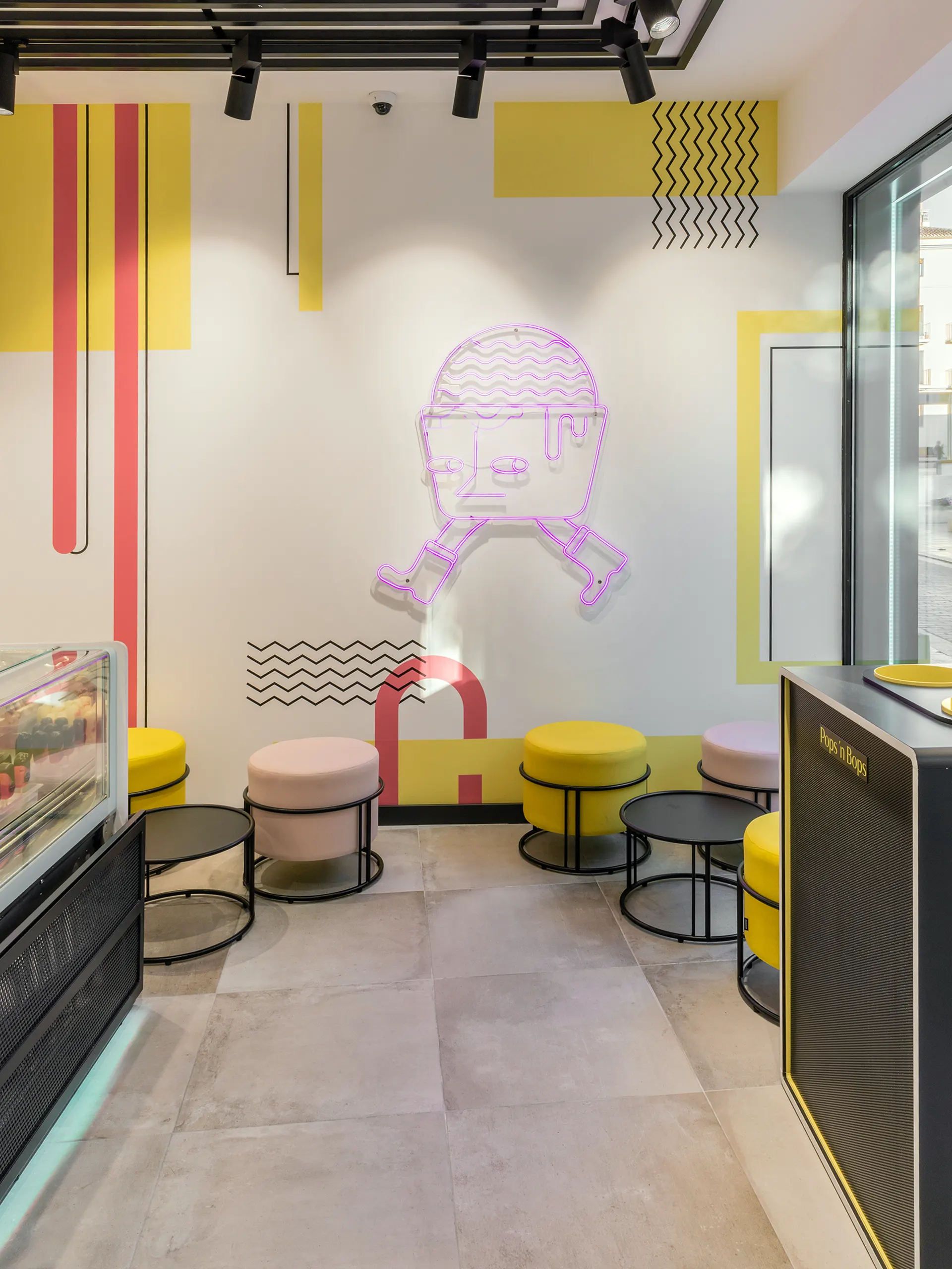 Pared decorativa en Pops'n Bops Valencia con formas geométricas de inspiración Memphis en amarillo y rojo, y un personaje de neón morado con forma de helado con piernas