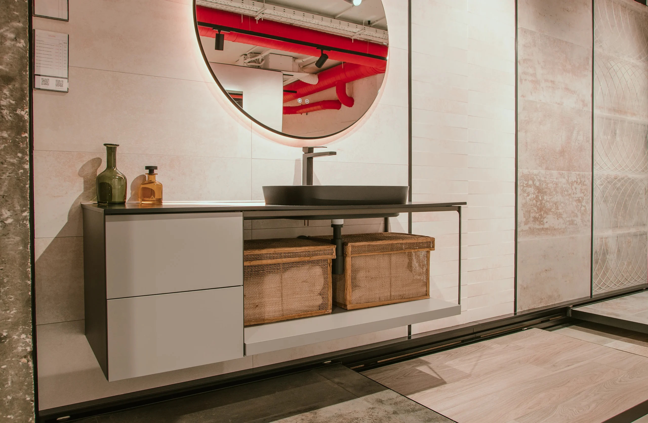 Mueble de baño suspendido con lavabo negro, espejo circular retroiluminado y cestos de fibra natural en la flagship de SALONI en Madrid