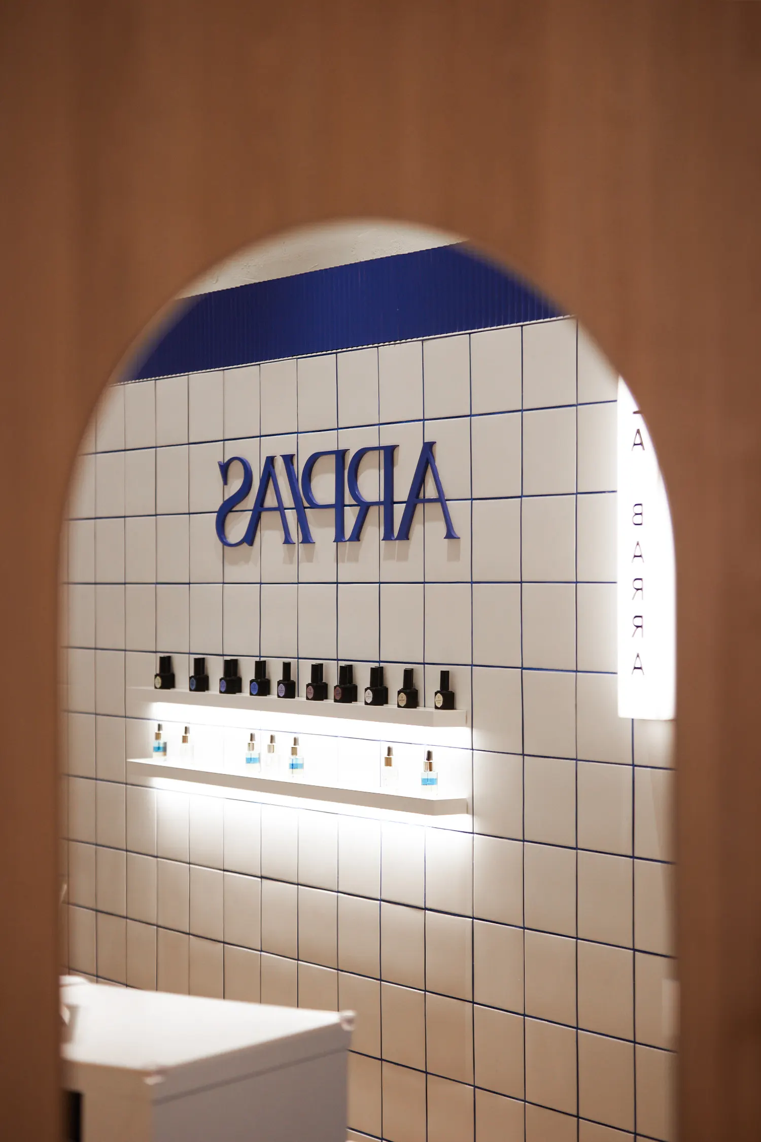 Logotipo de Arpías en letras azules sobre azulejo blanco con juntas azules, con vista a la barra de manicura y estantería de esmaltes, centro diseñado por huuun