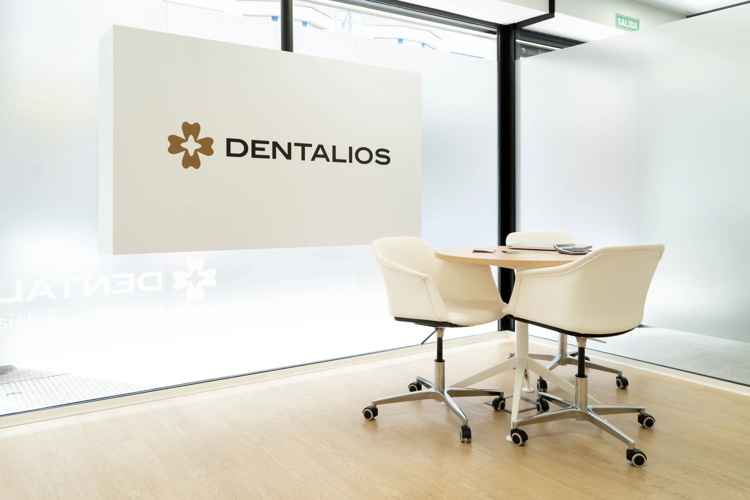 Panel suspendido con el logotipo de Dentalios sobre cristal esmerilado, junto a una mesa redonda con sillones blancos