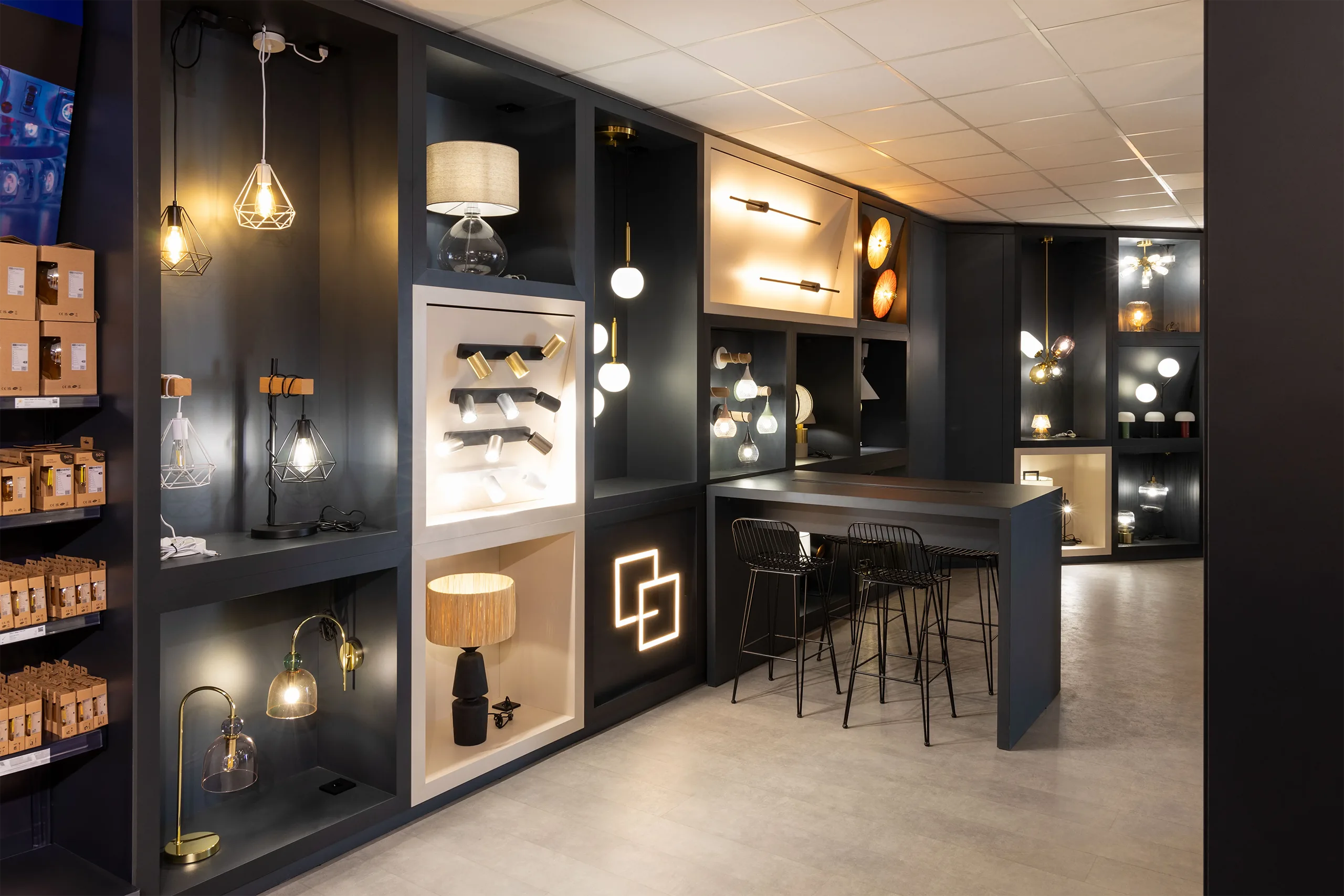 Estantería modular en negro con lámparas de mesa, apliques y colgantes expuestos en efectoLED Catarroja, con el icono de marca retroiluminado en la pared