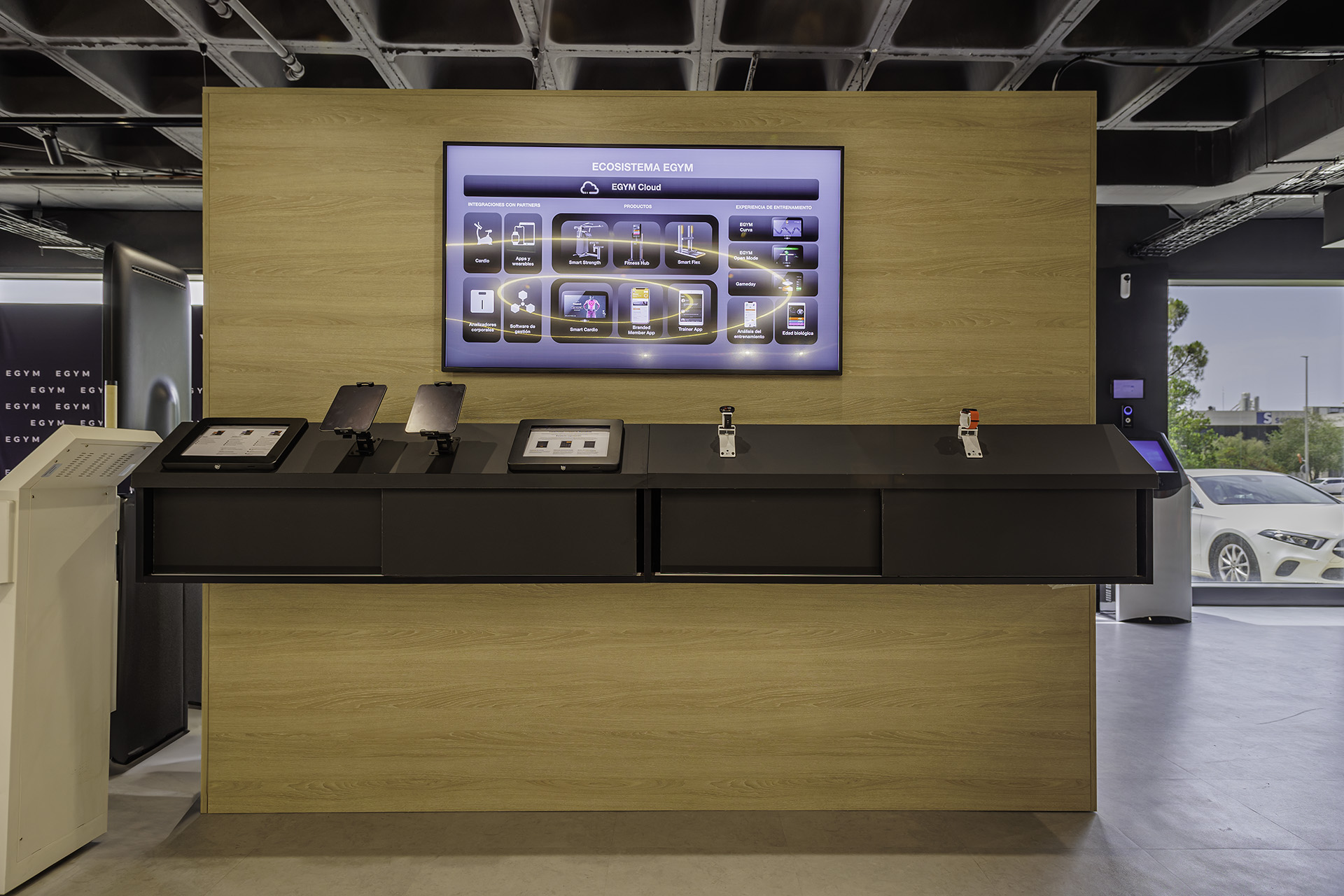 Pantalla con el esquema del ecosistema EGYM —EGYM Cloud, productos y experiencia de entrenamiento— sobre mostrador de madera con tablets y dispositivos wearables en exposición