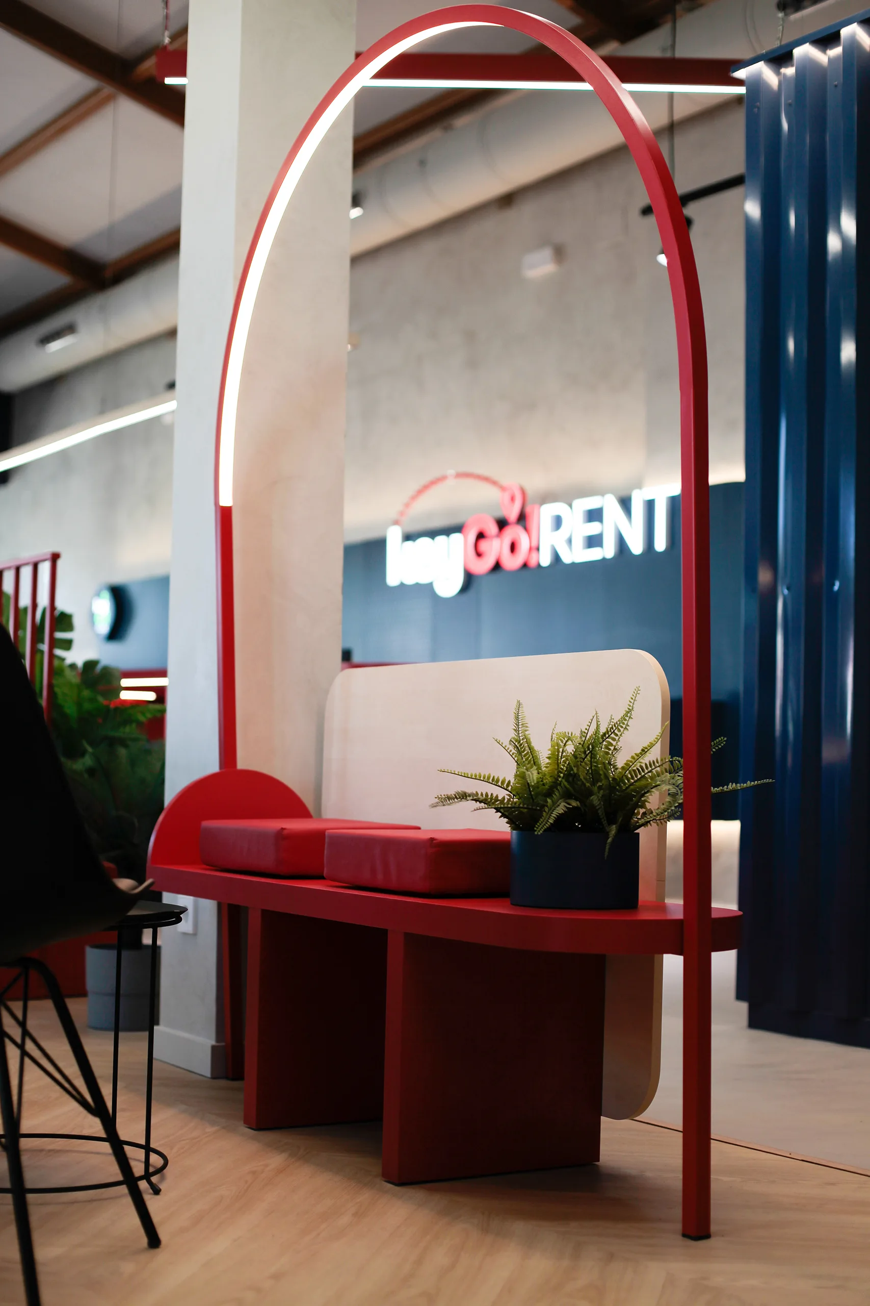 Banco curvo rojo con cojines bajo un arco luminoso, junto al rótulo retroiluminado de KeyGo Rent en Alicante