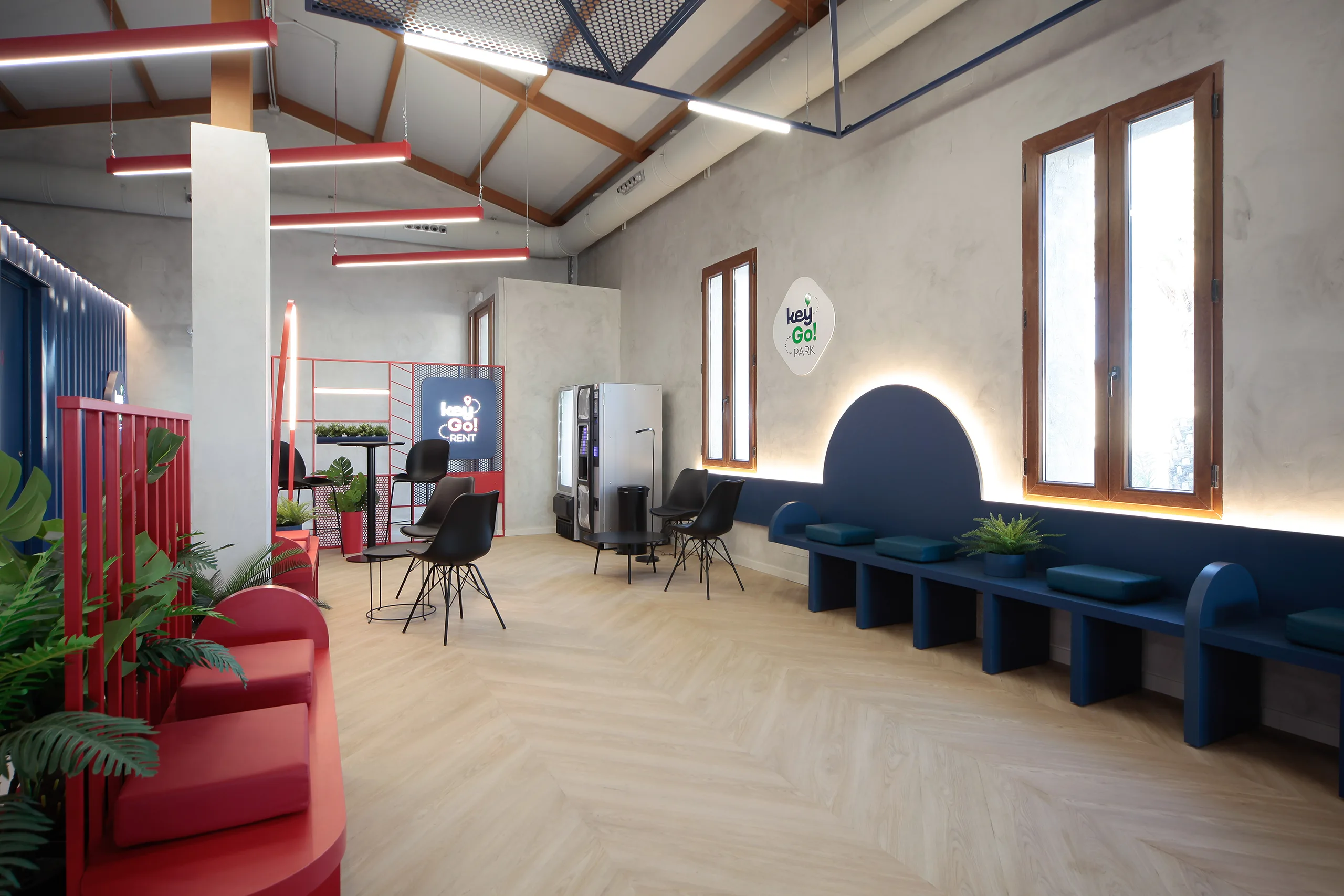 Sala de espera de KeyGo Rent con banco corrido azul petróleo bajo un arco luminoso, sillas negras y estructura roja de celosía, oficina de alquiler de coches en Alicante