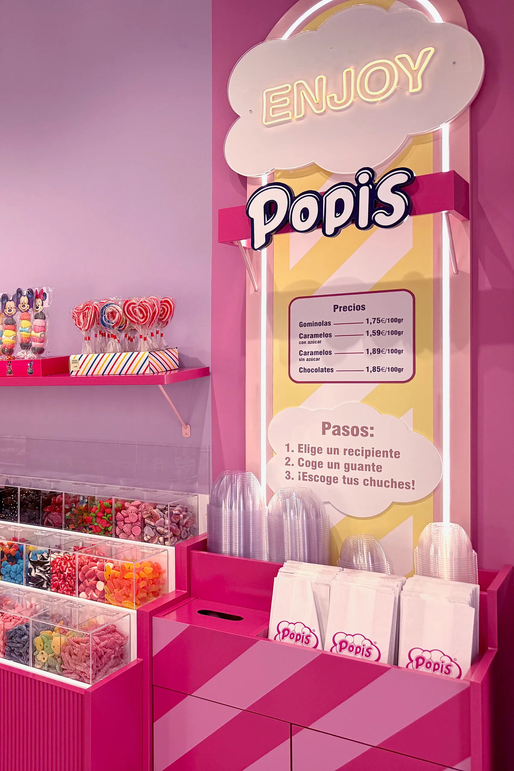 Cartel en forma de nube con el neón «Enjoy Popis», lista de precios y pasos para servirse chuches a granel, junto al mostrador rosa de POPIS Valencia