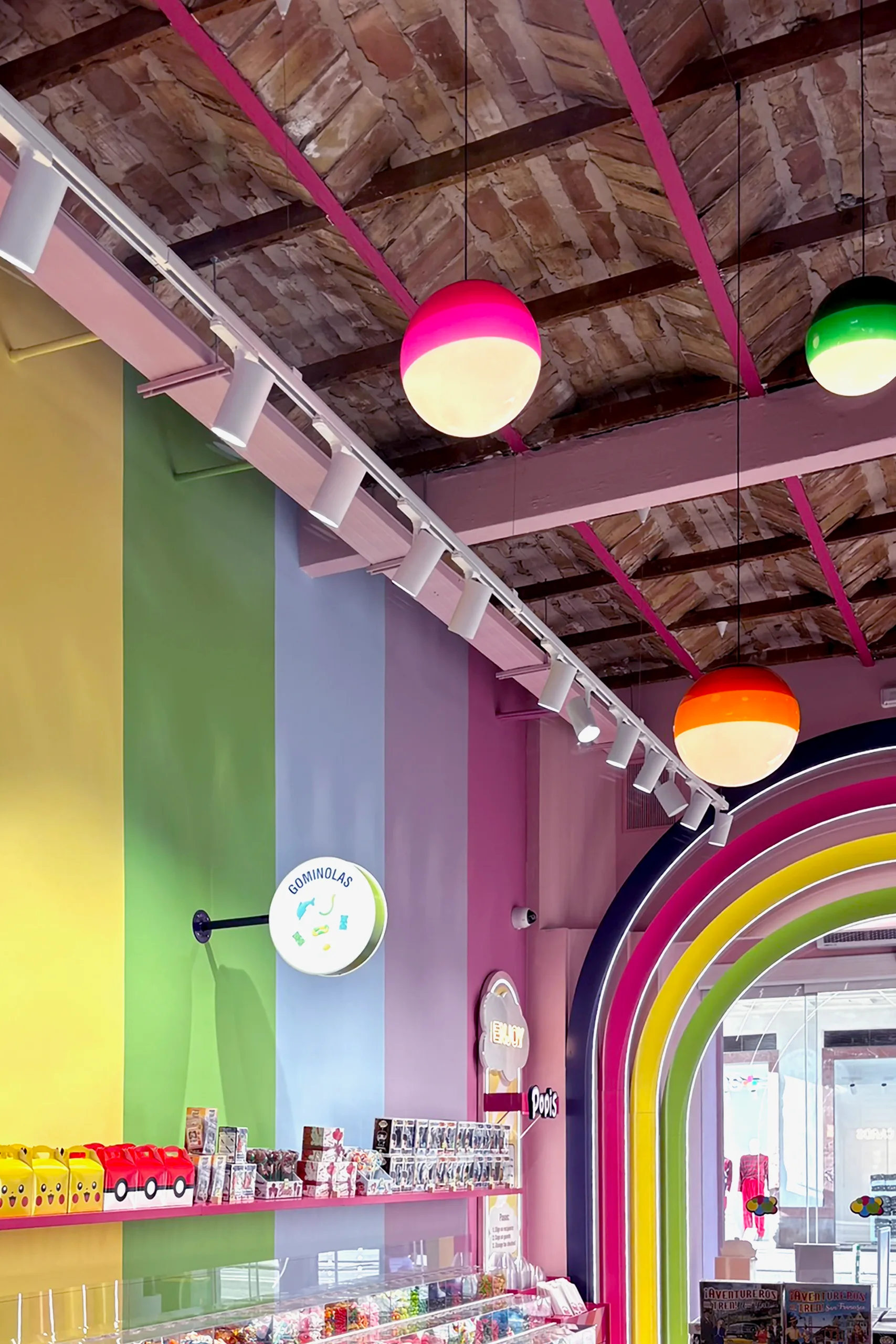 Techo original de bovedilla cerámica con raíl de iluminación rosa y lámparas esféricas colgantes, sobre la pared arcoíris y el cartel «Gominolas» de POPIS Valencia