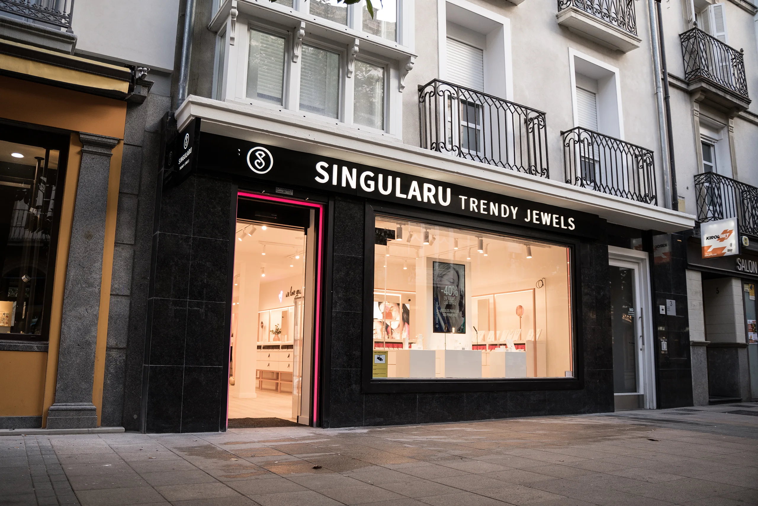 Fachada de Singularu Trendy Jewels en un edificio con balcones de forja, puerta con neón rosa y escaparate iluminado