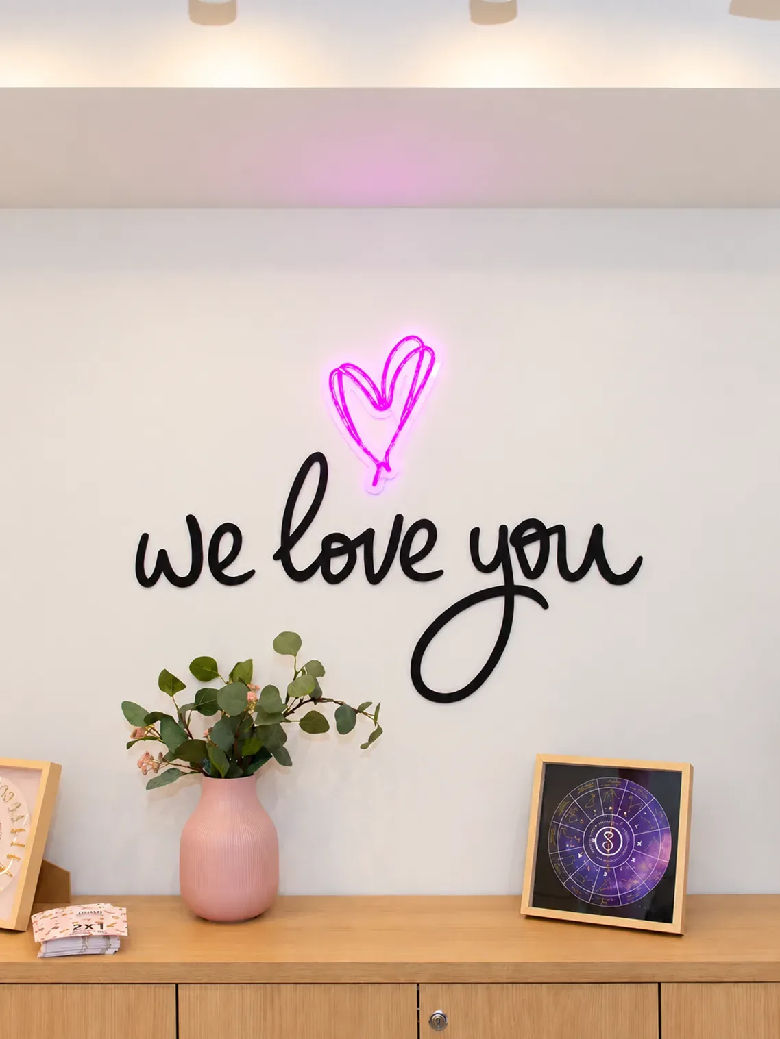 Neón rosa con corazón y rótulo «we love you» sobre un mueble de roble con jarrón rosa en Singularu
