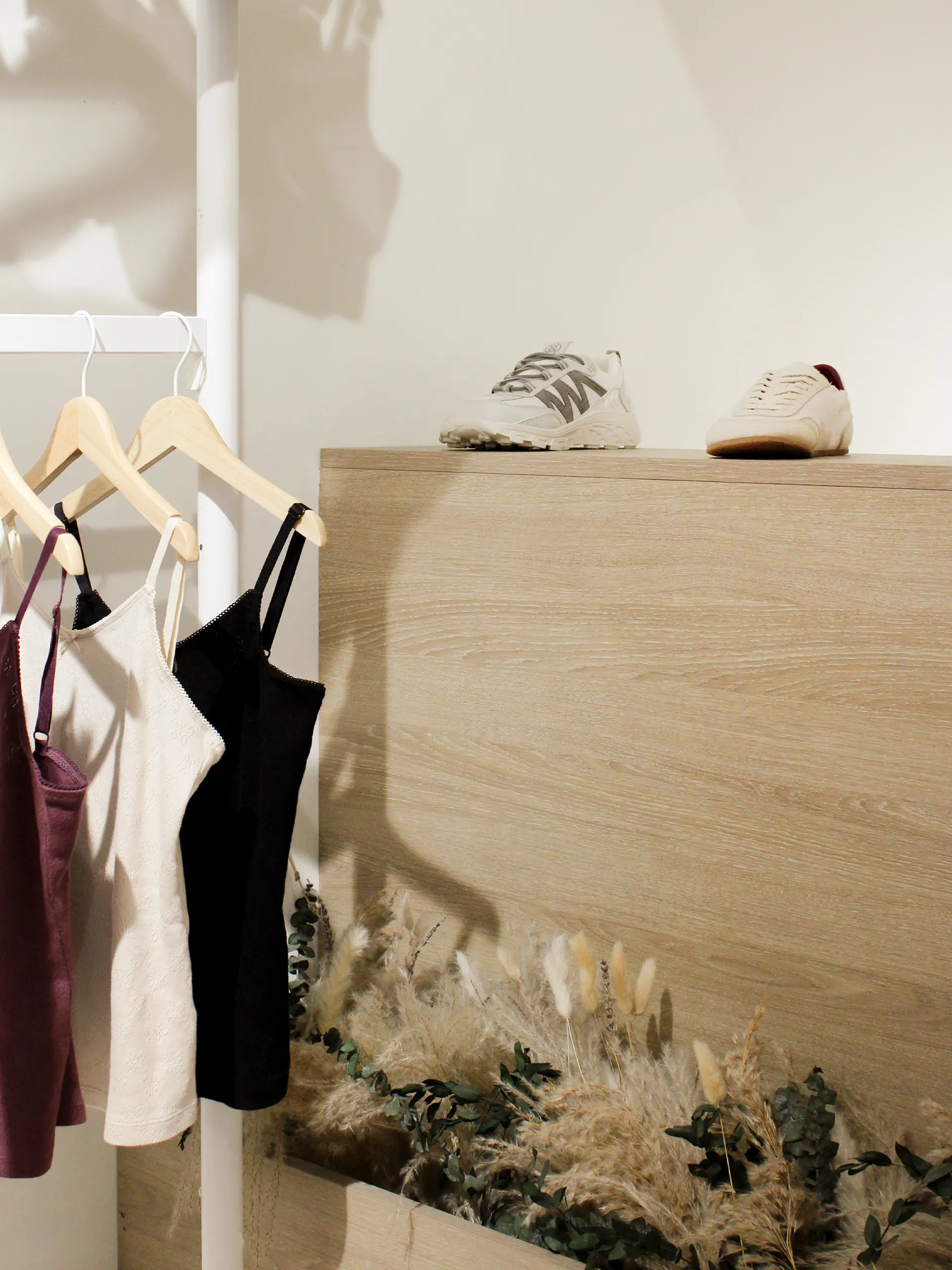 Tops de tirantes en perchas de madera junto a un mueble de madera clara con zapatillas y jardinera de flores secas y eucalipto