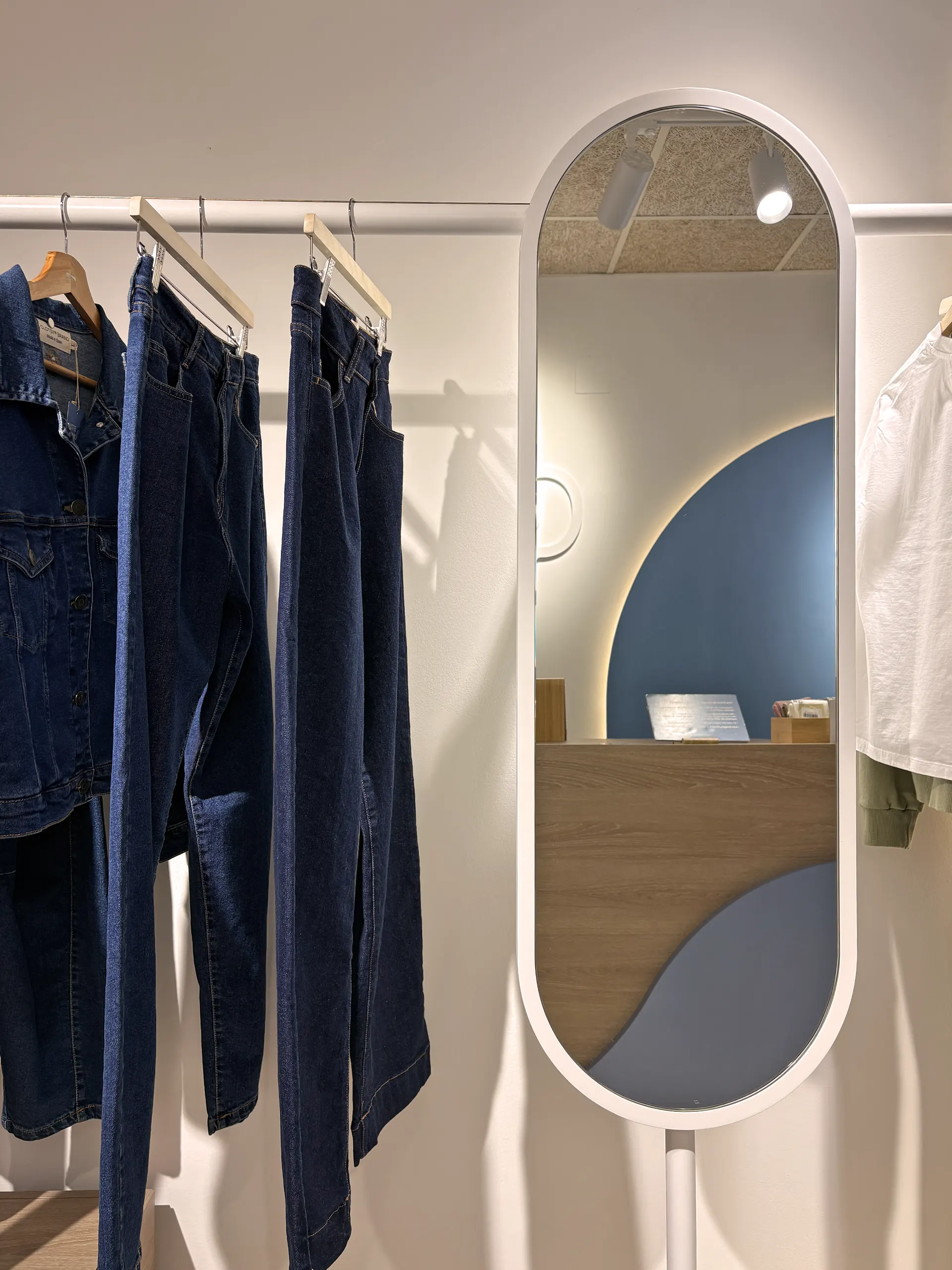 Espejo ovalado de marco blanco junto a una barra con chaqueta y vaqueros, que refleja el mostrador de madera y el arco azul retroiluminado
