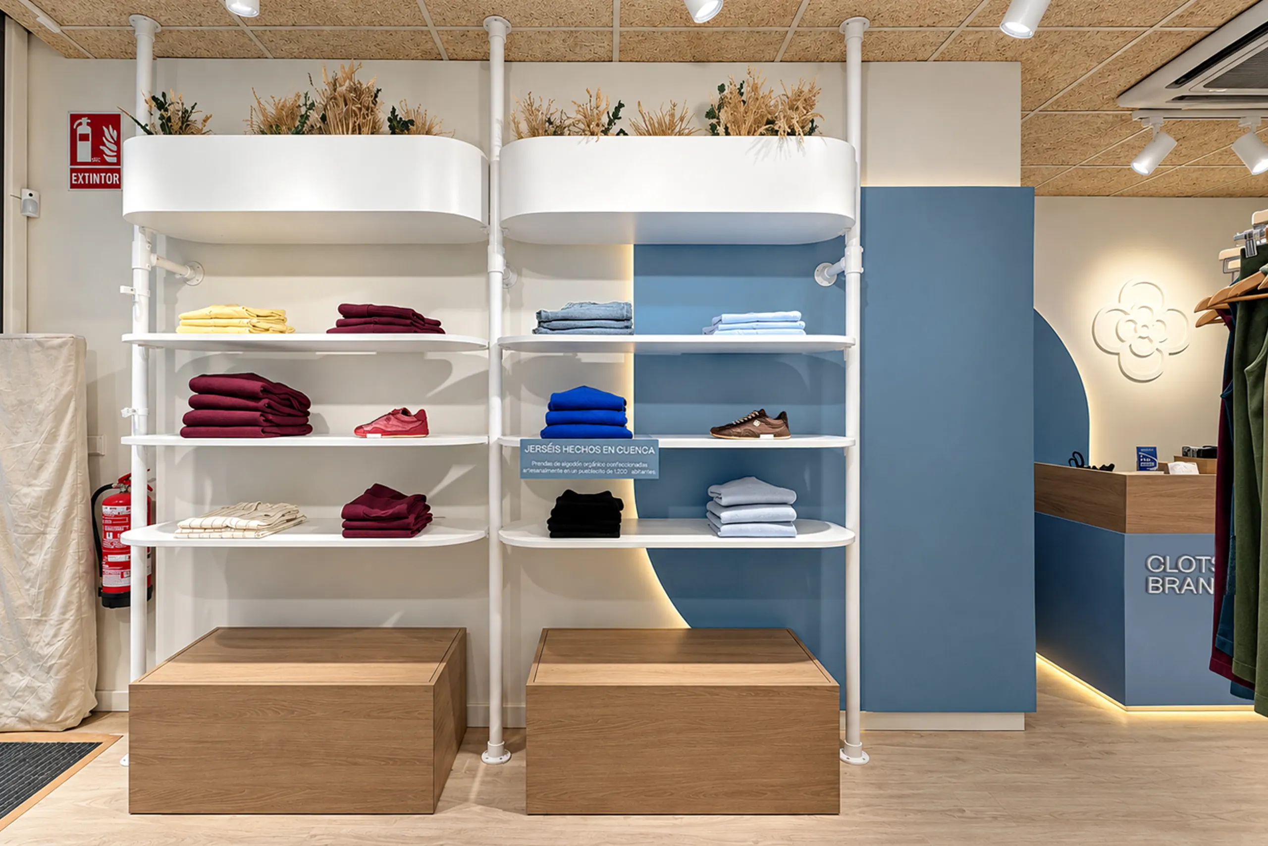 Estanterías blancas de baldas curvas con jardineras de espigas secas, prendas dobladas, zapatillas y peanas de madera frente a un arco azul retroiluminado