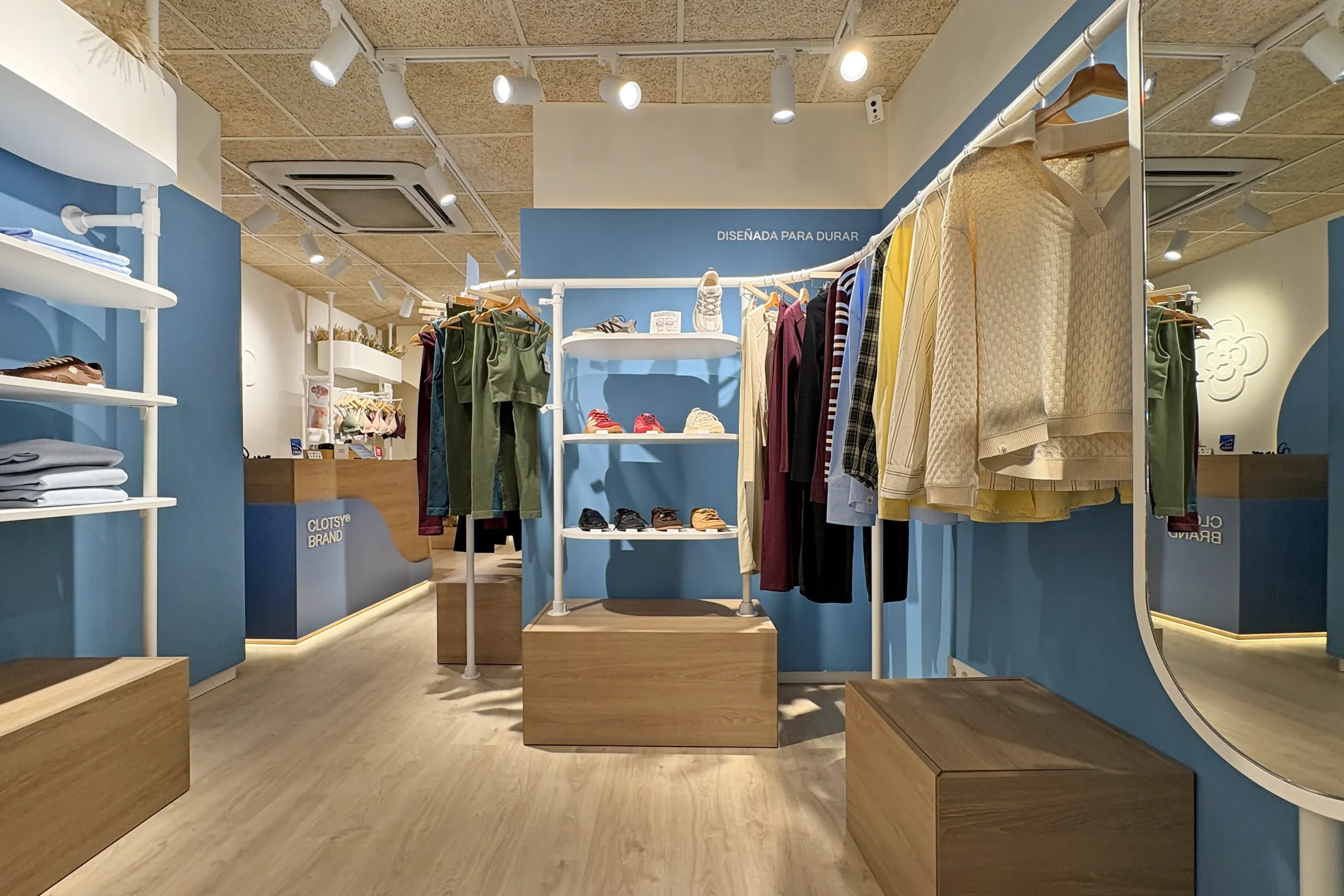 Estanterías blancas de baldas curvas con jardineras de espigas secas, prendas dobladas, zapatillas y peanas de madera frente a un arco azul retroiluminado