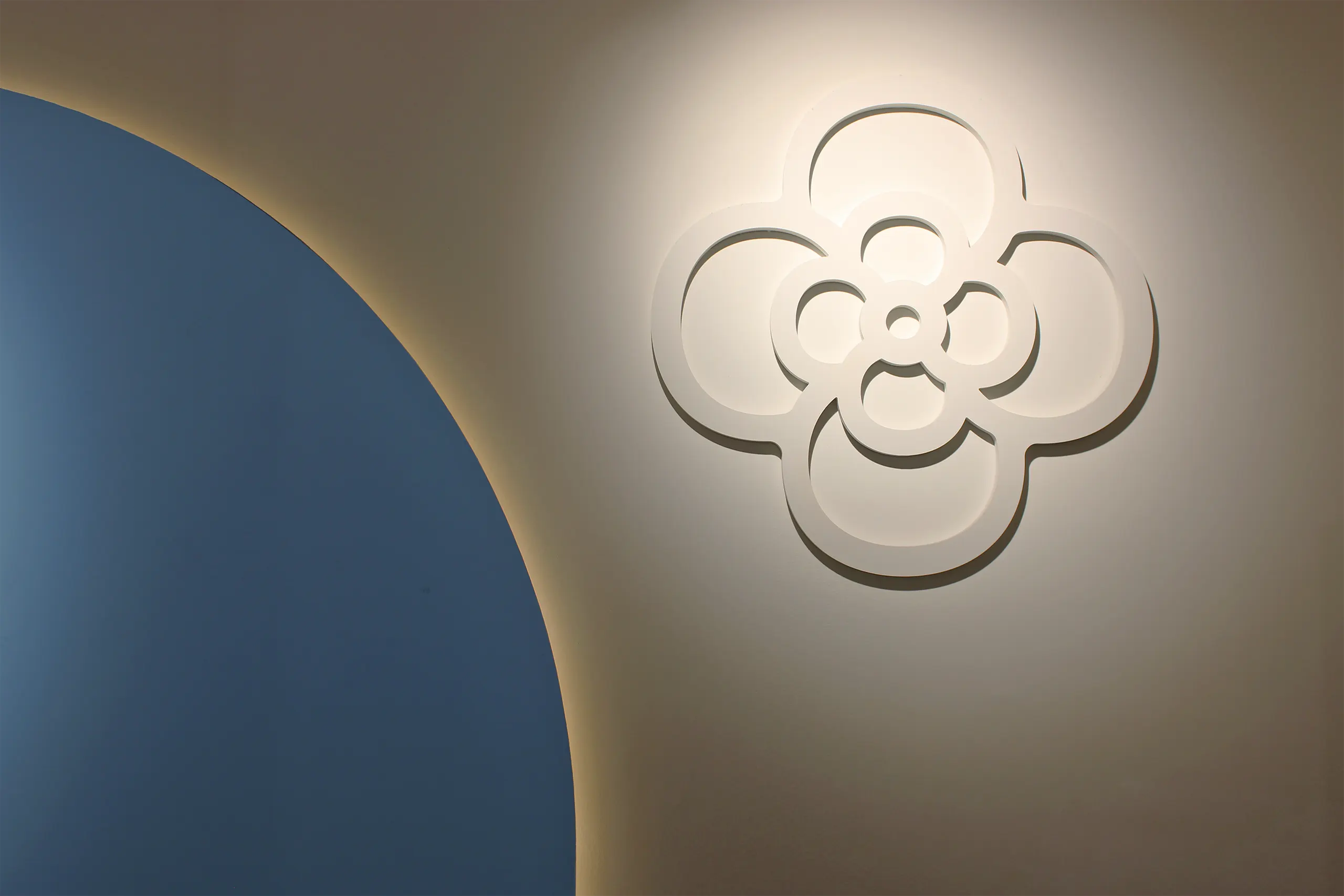 Isotipo floral de Clotsy en relieve blanco iluminado sobre pared clara, junto a un arco azul con luz indirecta