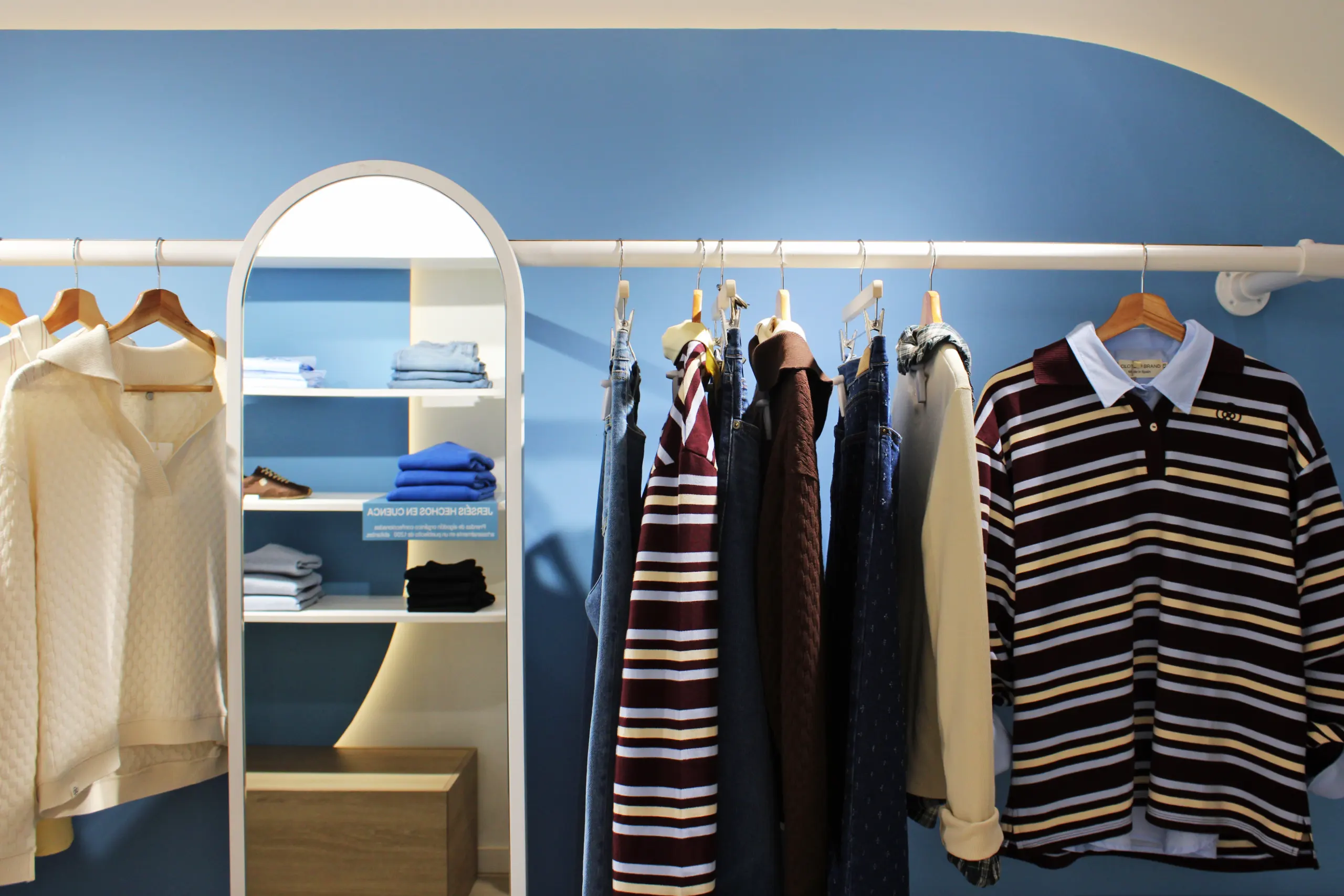 Pared azul de forma orgánica con barra blanca de prendas colgadas y una estantería en arco con ropa doblada, zapatillas y peana de madera