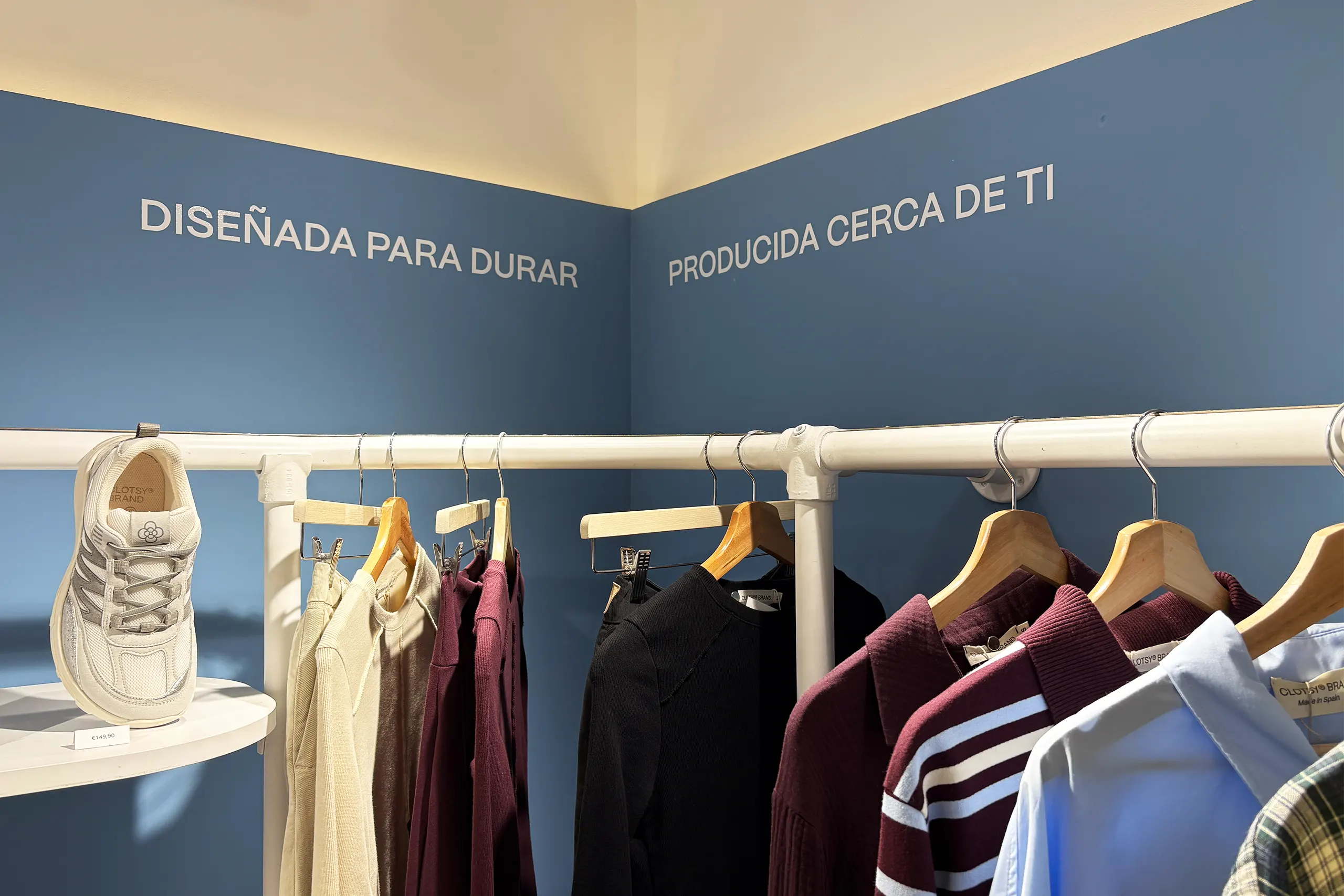 Rincón de pared azul con los mensajes «Diseñada para durar» y «Producida cerca de ti» sobre una barra blanca con prendas y una zapatilla