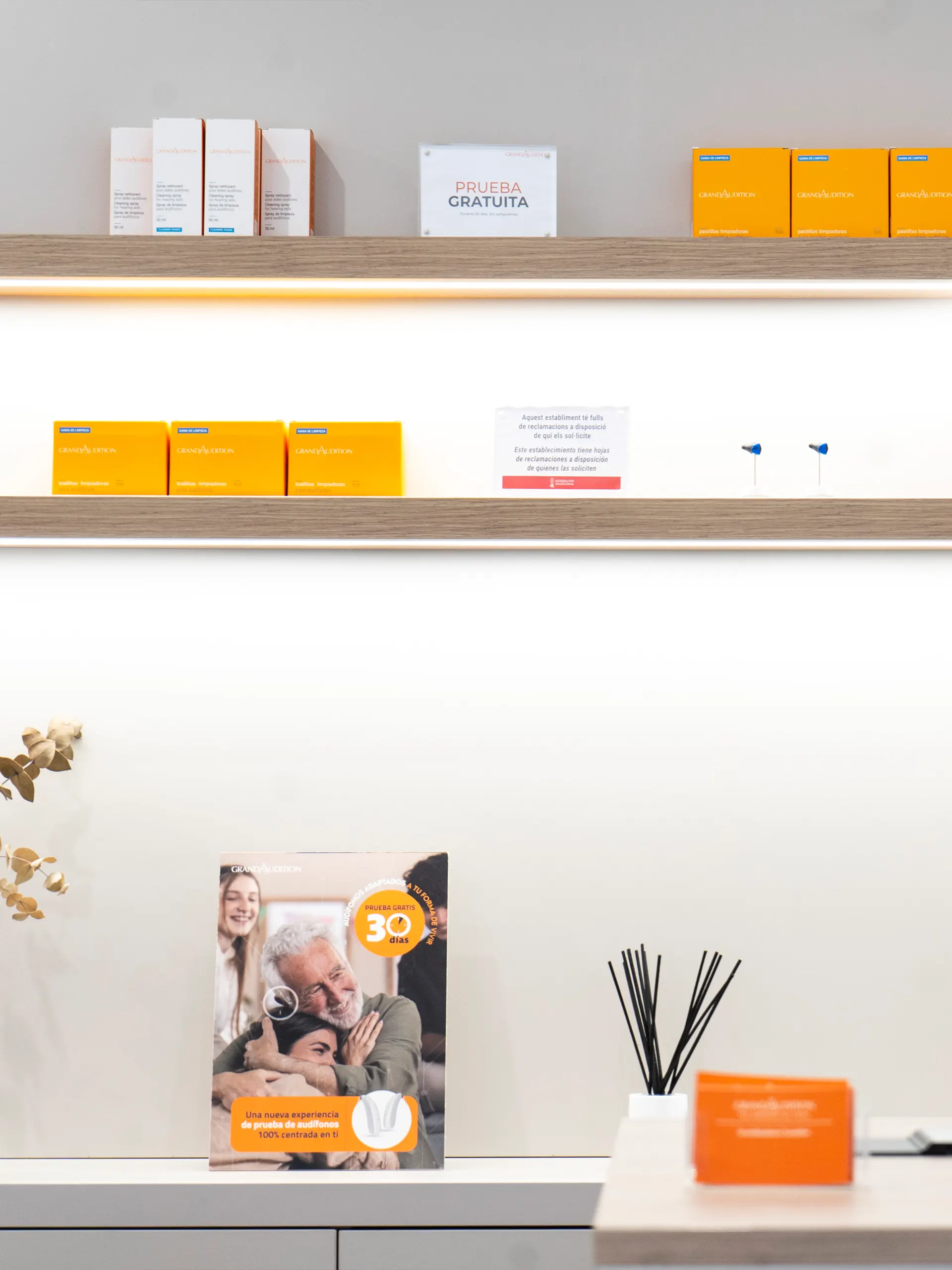 Estantes de madera con iluminación LED y productos de limpieza de audífonos en naranja de GrandAudition, con folleto promocional sobre el mueble