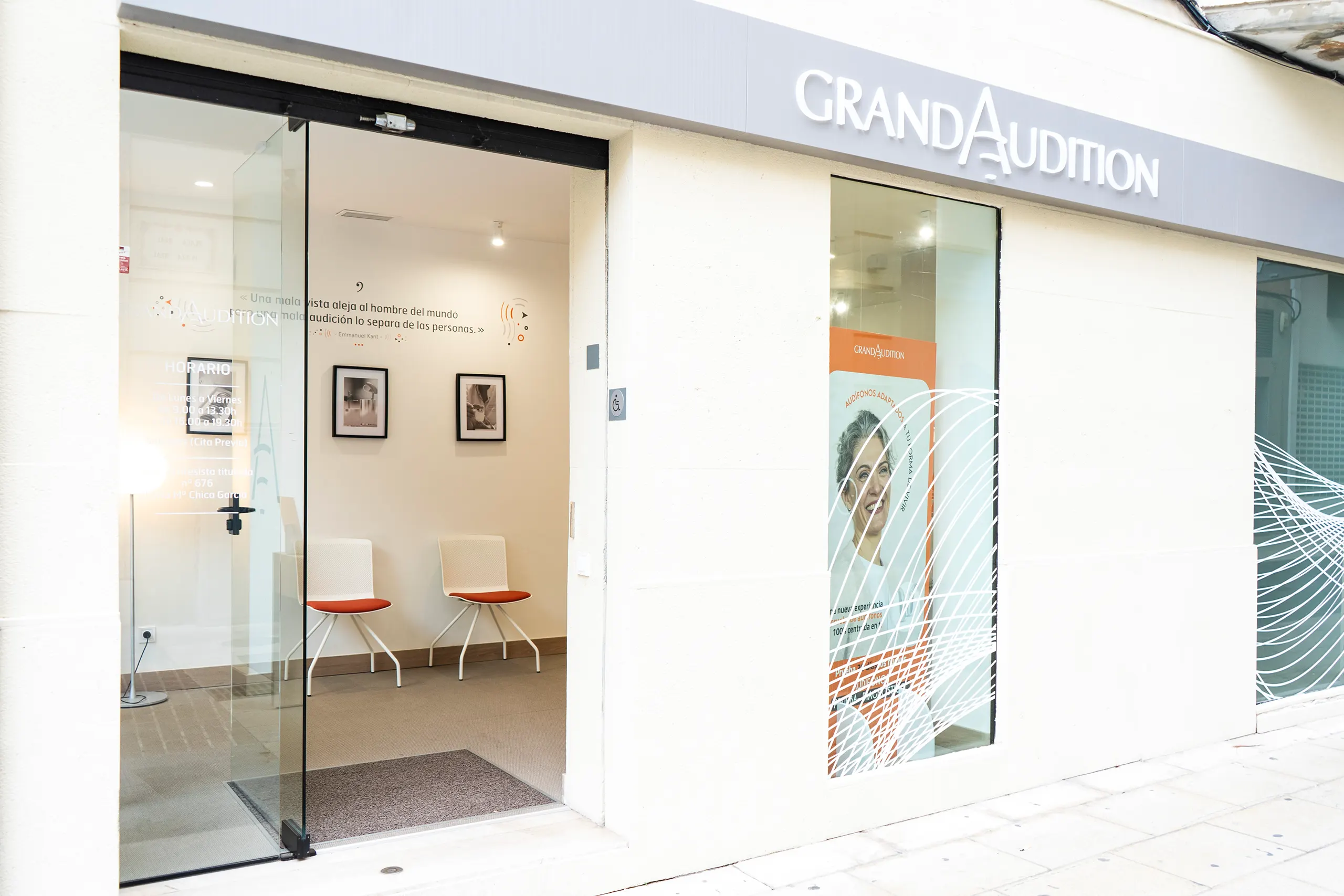 Entrada de GrandAudition con puerta de vidrio abierta hacia la sala de espera con sillas blancas y asientos naranjas