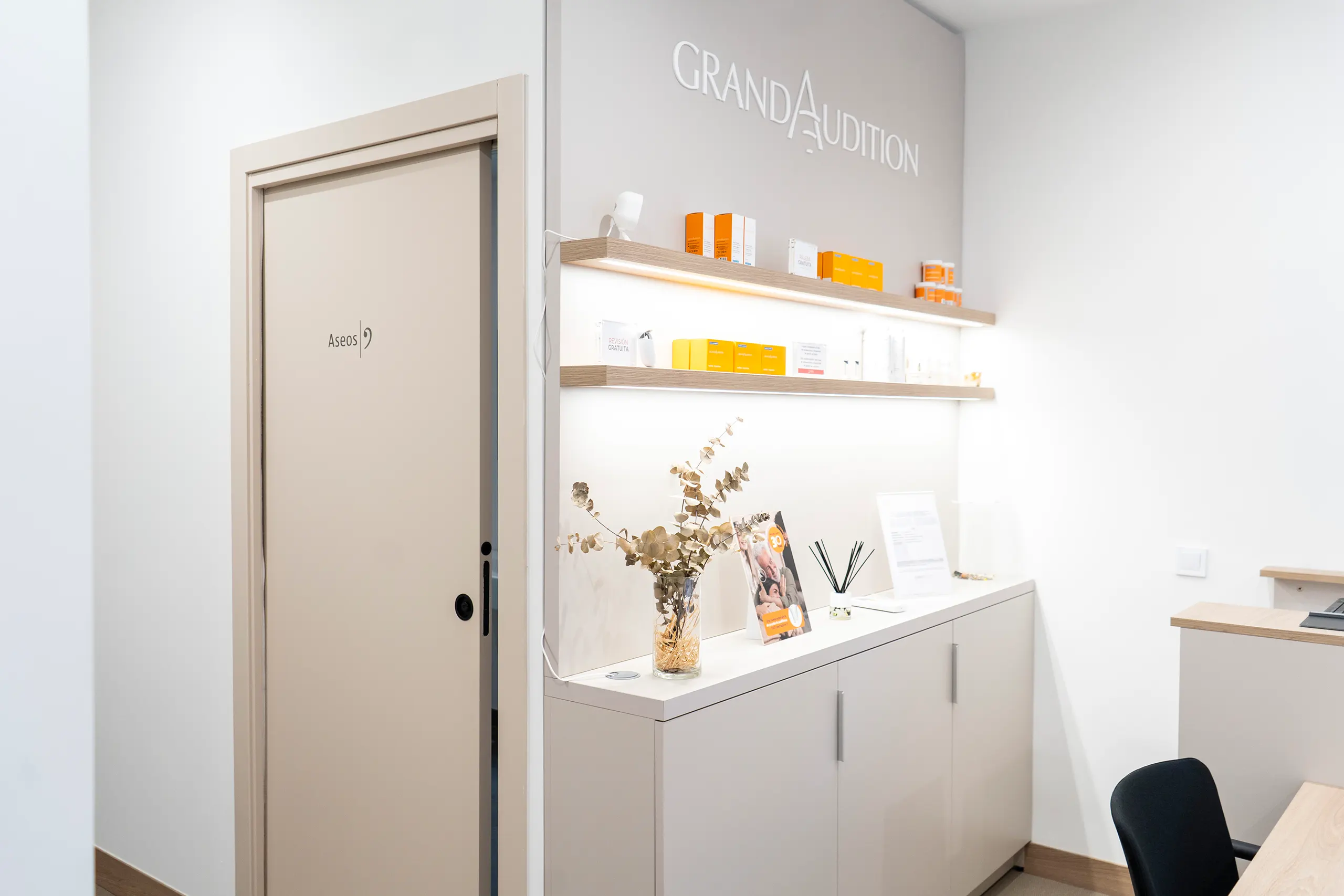 Mueble expositor con estantes de madera iluminados y productos naranjas de GrandAudition junto a la puerta de los aseos