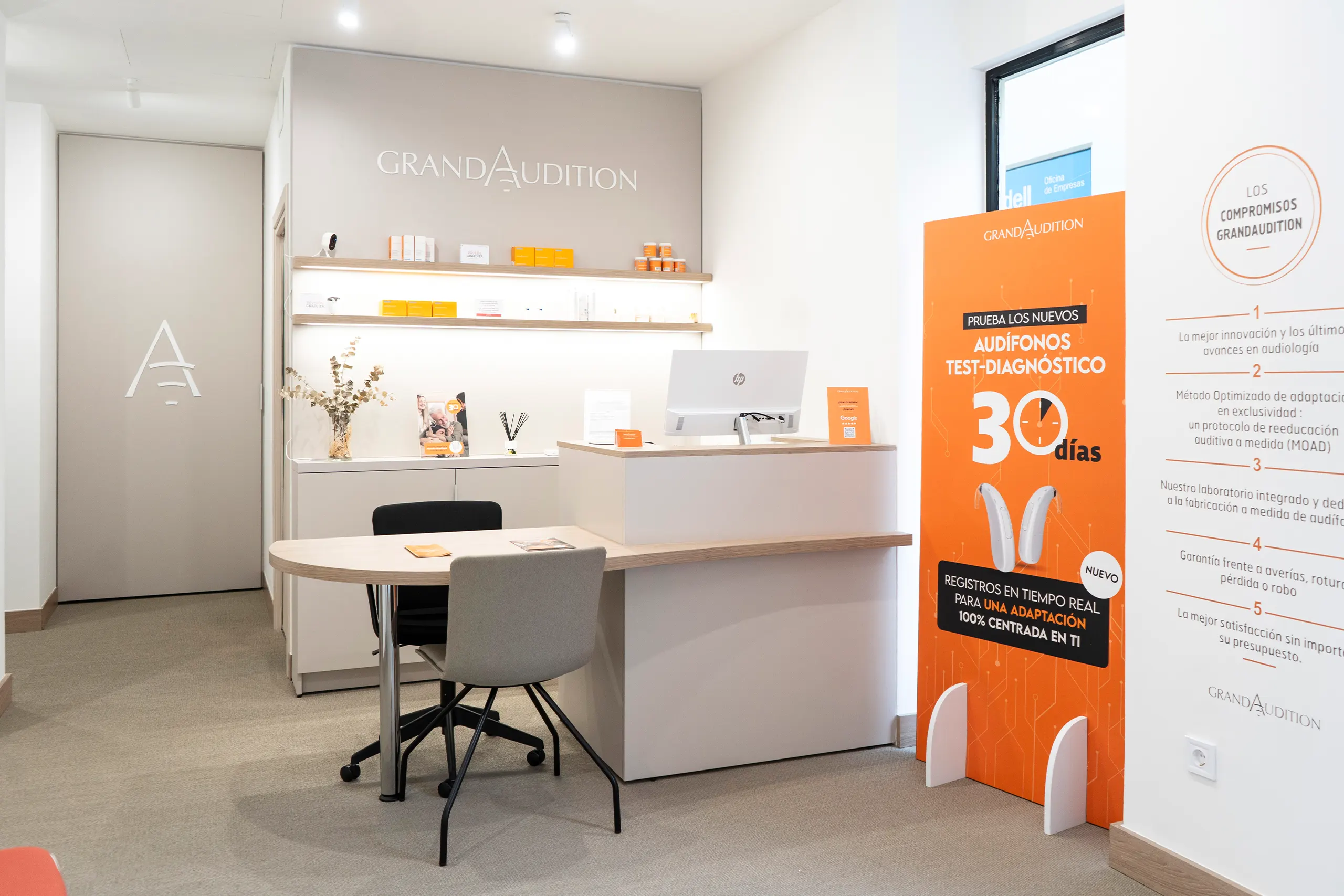 Zona de recepción de GrandAudition con mesa de atención en madera, tótem naranja de campaña y gráfica mural con los compromisos de la marca