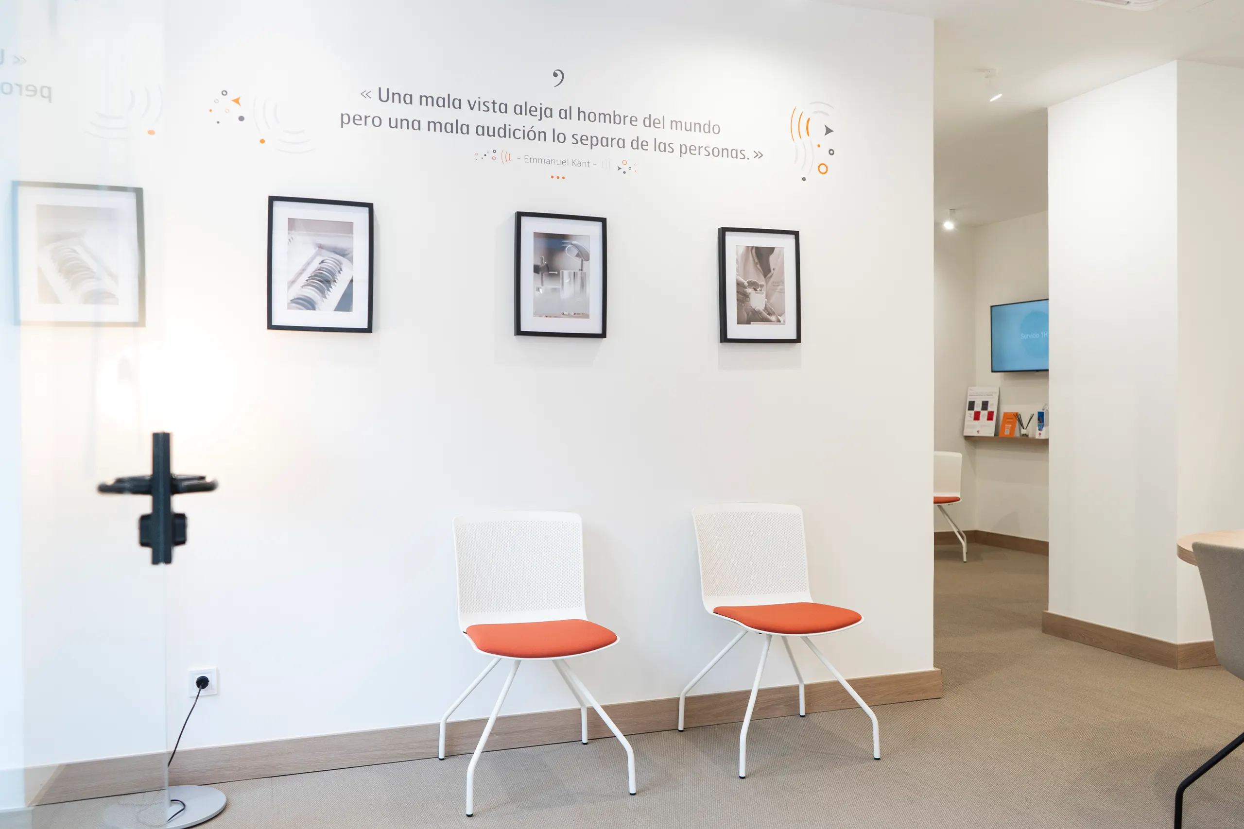 Sala de espera de GrandAudition con dos sillas blancas de asiento naranja, fotografías enmarcadas y una cita de Kant sobre la audición en la pared