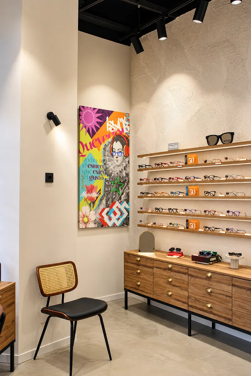 Lienzo de estética pop junto a una pared de monturas de colores sobre baldas iluminadas y una silla de rejilla en la óptica Quevedos