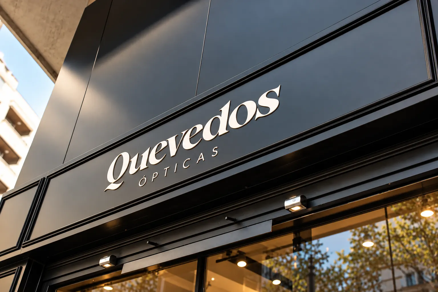 Rótulo «Quevedos Ópticas» en letras blancas sobre la fachada negra con molduras de la óptica en Gandía